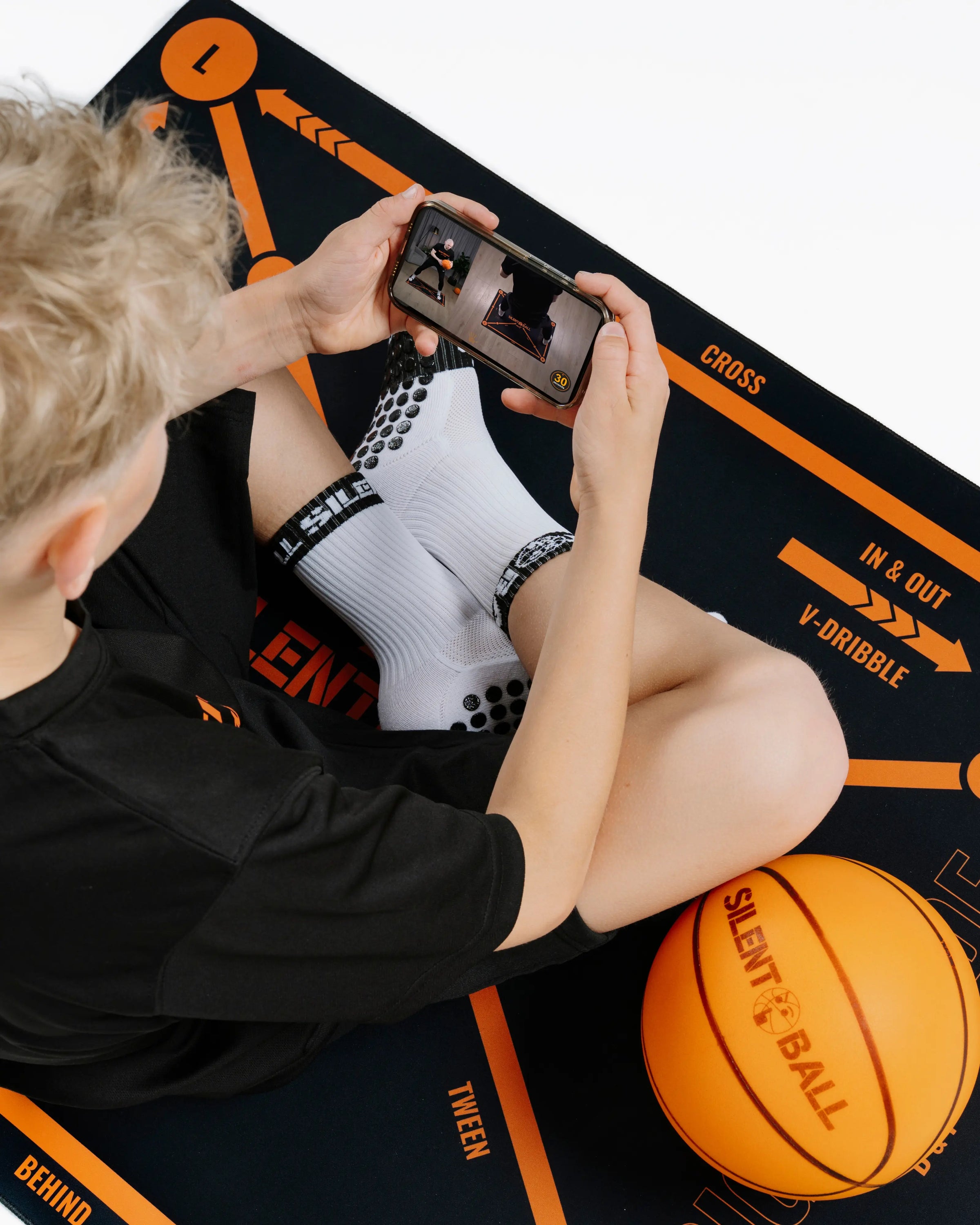 A client sitting comfortably on a training mat, focused on a smartphone or tablet displaying a SilentBall Academy video training session from the app